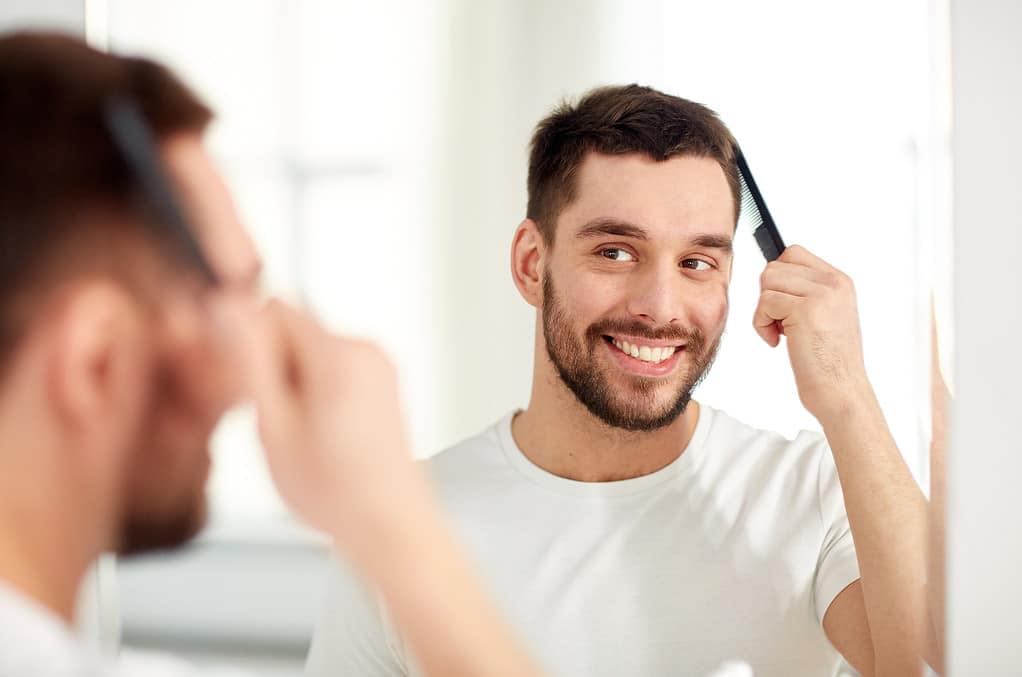 beauty, grooming and people concept - smiling young man looking to mirror and brushing hair with comb at home bathroom