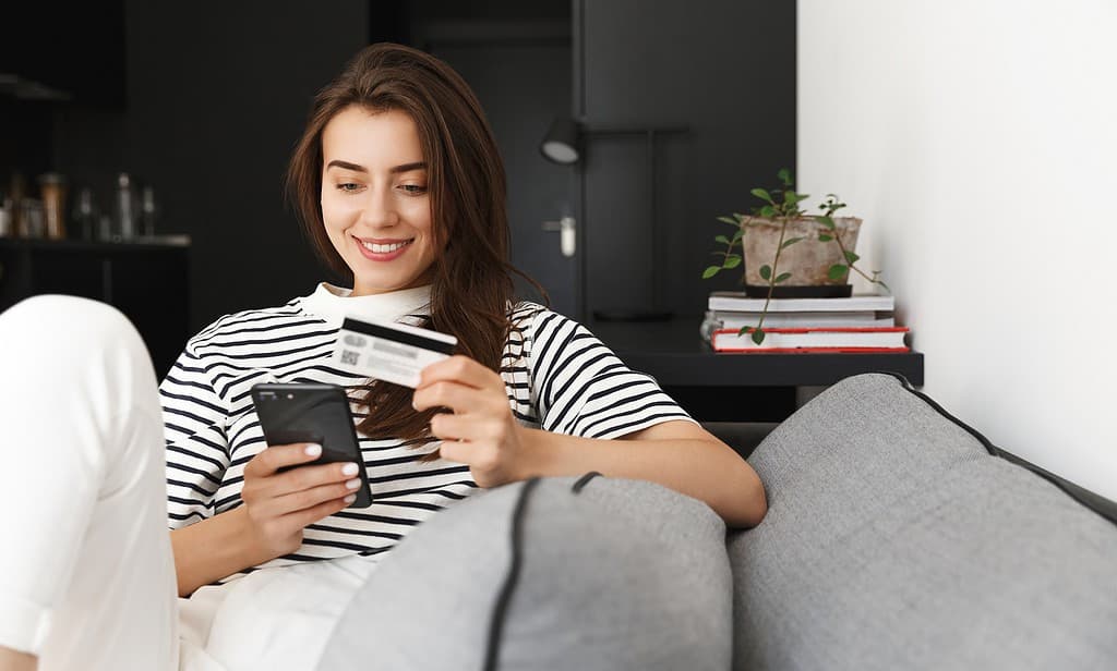 Portrait of resting girl paying on mobile phone for shopping online, holding credit card and holding smartphone, lying on sofa at home and paying in internet.